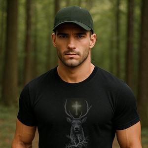 May include: Black crew neck t-shirt featuring a deer head with antlers and a cross design. The graphic is in shades of gray and white. The person in the image is wearing a green baseball cap and khaki pants.