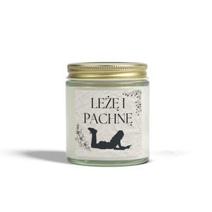 Könnte beinhalten: Eine weiße Kerze in einem Glasgefäß mit goldenem Deckel. Das Etikett auf dem Gefäß zeigt eine schwarze Silhouette einer Person, die ein Buch liest, und den Text "LEŽE I PACHNE".