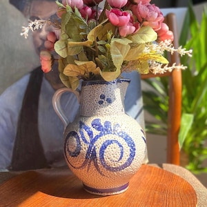 May include: A ceramic pitcher with a speckled beige glaze and a blue hand-painted floral swirl design. The pitcher features a curved handle, a wide rounded body, and a flared rim, filled with a bouquet of pink artificial flowers and green leaves.