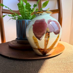 May include: A small, round ceramic vase with a cream-colored base, featuring hand-painted dark red floral shapes and brown vertical brushstrokes. The vase has a narrow, dark-rimmed opening and a smooth, glossy finish with visible glaze variations.