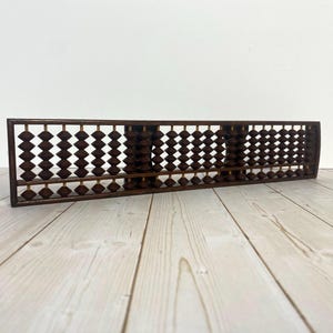 May include: A vintage wooden abacus with dark brown beads. The abacus is rectangular, with multiple rows of beads separated by wooden dividers. The beads are diamond-shaped and appear to be made of a dark wood. The abacus is set on a light-colored wooden surface.