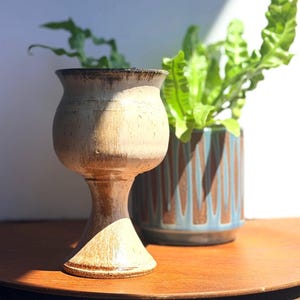 May include: A ceramic goblet with a flared rim and a pedestal base, featuring a cream-colored glaze with brown speckles and dark brown accents along the rim and base. The surface has a textured appearance with visible variations in the glaze color.