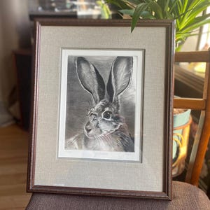 May include: Framed black and white print of a hare with long ears. The artwork is in a brown wooden frame with a light gray mat. The hare is detailed with shading and texture, giving it a realistic appearance. The print is signed and numbered.