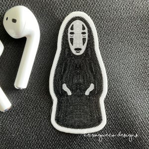 May include: A white sticker featuring a character from Spirited Away, with a black body and a white face. The sticker is next to a white wireless earbud. The sticker has the text "krisoguecco designs" in cursive.