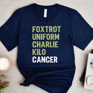 May include: Navy blue t-shirt with the words "FOXTROT UNIFORM CHARLIE KILO CANCER" printed in green and white. The shirt is folded and placed on a light-colored surface, with a candle, book, and glasses nearby.