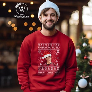 Könnte beinhalten: Roter Weihnachtspullover mit dem Text "GARNACHO HO HO" und "MERRY CHRISTMAS" in Weiß. Der Pullover zeigt eine Grafik einer Person mit Weihnachtsmütze. Ein festlicher Feiertagsartikel.