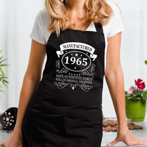 May include: A black apron with white text that reads "Manufactured 1965 100% Authentic Parts Still in Original Wrapping". The apron has a white star and a white banner with a decorative design.