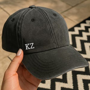 Peut inclure: Casquette de baseball gris foncé avec les lettres "KZ" brodées en blanc sur le devant. La casquette est faite d'une matière douce et usée et possède une visière incurvée. La casquette est tenue par une personne.
