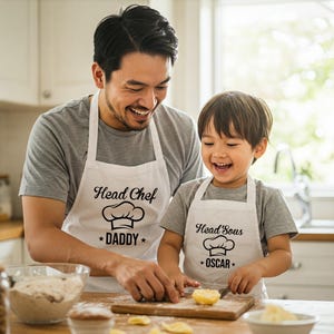 May include: A father and son wearing white aprons with the text "Head Chef *Daddy*" and "Head Sous *Oscar*" while cooking in the kitchen.