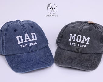 May include: Two denim baseball caps, one blue and one gray, with white embroidered text. The blue cap says "DAD EST. 2025" and the gray cap says "MOM EST. 2025".