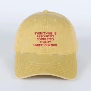 May include: Mustard yellow baseball cap with the embroidered text "EVERYTHING IS ABSOLUTELY COMPLETELY TOTALLY UNDER CONTROL" in red. The cap has a curved brim and a classic six-panel design.