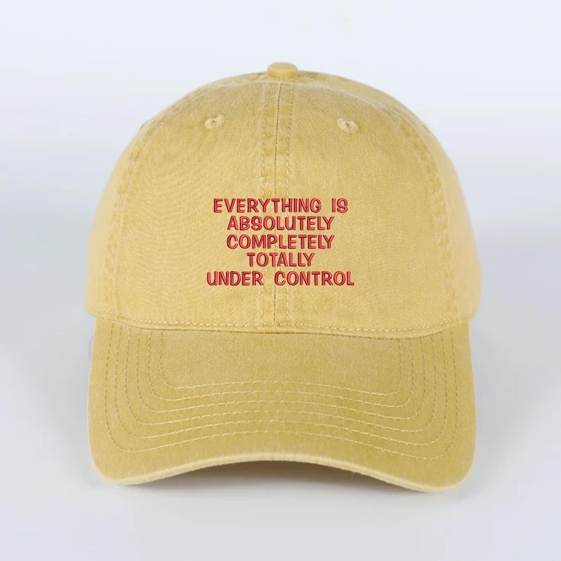 May include: Mustard yellow baseball cap with the embroidered text "EVERYTHING IS ABSOLUTELY COMPLETELY TOTALLY UNDER CONTROL" in red. The cap has a curved brim and a classic six-panel design.