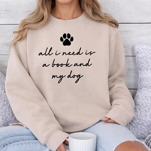 May include: Beige sweatshirt with a black paw print and the text "all i need is a book and my dog". The sweatshirt is paired with blue jeans. A white mug is held in the hands.
