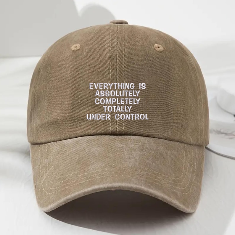 May include: A khaki-colored baseball cap with the phrase "EVERYTHING IS ABSOLUTELY COMPLETELY TOTALLY UNDER CONTROL" embroidered in white. The cap has a curved brim and a button on top.