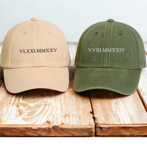 Może przedstawiać: Dwie czapki bejsbolówki, jedna beżowa i jedna oliwkowa, są pokazane na drewnianej powierzchni. Beżowa czapka ma wydrukowaną rzymską datę VI.XXI.MMXXV. Oliwkowa czapka ma wydrukowaną rzymską datę V.VIII.MMXXIV.