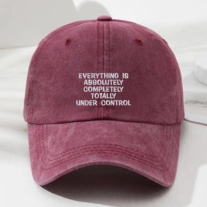 May include: A burgundy baseball cap with a curved brim and a top button. The cap features the white text "EVERYTHING IS ABSOLUTELY COMPLETELY TOTALLY UNDER CONTROL" across the front. The cap is made of a soft, textured material.