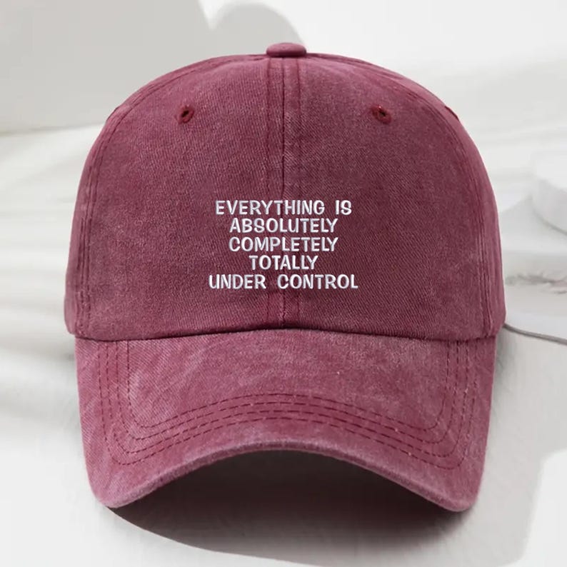May include: A burgundy baseball cap with a curved brim and a top button. The cap features the white text "EVERYTHING IS ABSOLUTELY COMPLETELY TOTALLY UNDER CONTROL" across the front. The cap is made of a soft, textured material.