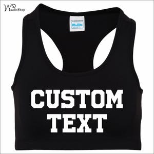 May include: Black sports bra with the words "CUSTOM TEXT" in bold white letters. The bra has a racerback design and a scoop neckline. A small white tag is visible near the neckline.