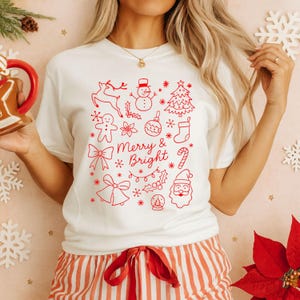 Könnte beinhalten: Weißes T-Shirt mit rotem Weihnachtsdesign. Das Design zeigt ein Rentier, einen Schneemann, einen Weihnachtsbaum, Lebkuchenmännchen, Ornamente und den Text "Merry & Bright". Das Shirt wird mit rot-weiß gestreiften Pyjamahosen kombiniert.