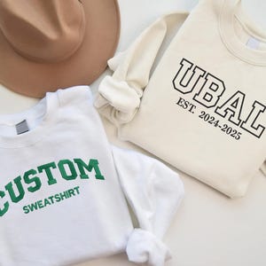 May include: Two white sweatshirts with different text printed on them. One sweatshirt says "CUSTOM SWEATSHIRT" in green letters. The other sweatshirt says "UBAL EST. 2024-2025" in black letters.