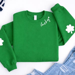 Puede incluir: Una sudadera verde con la palabra "Lucky" en escritura blanca y un trébol blanco. Un trébol blanco también está en la manga izquierda. La sudadera está doblada sobre una superficie blanca con jeans azules y un sombrero a rayas.