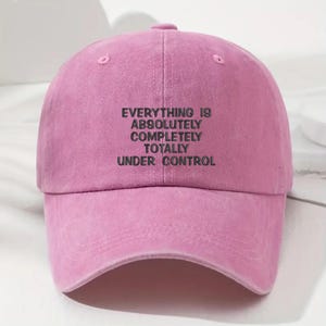 May include: A pink baseball cap with the embroidered text "EVERYTHING IS ABSOLUTELY COMPLETELY TOTALLY UNDER CONTROL" in black. The cap has a curved brim and a button on top.