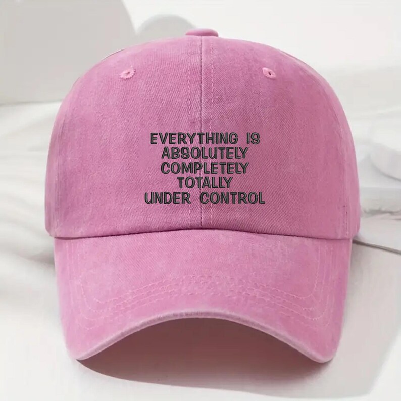 May include: A pink baseball cap with the embroidered text "EVERYTHING IS ABSOLUTELY COMPLETELY TOTALLY UNDER CONTROL" in black. The cap has a curved brim and a button on top.