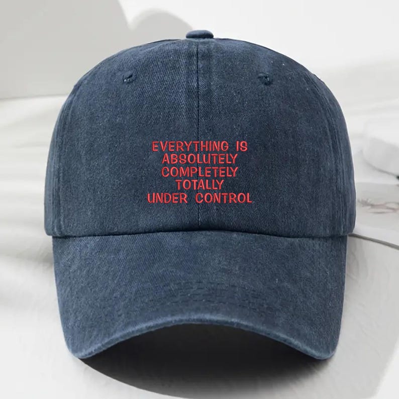 May include: A navy blue baseball cap with the embroidered text "EVERYTHING IS ABSOLUTELY COMPLETELY TOTALLY UNDER CONTROL" in red. The cap has a curved brim and a button on top.