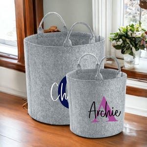 Op de afbeelding: Twee grijze opbergmanden van vilt met handvatten. De grotere mand heeft de naam "Charlie" in witte letters met een donkerblauwe cirkel erachter. De kleinere mand heeft de naam "Archie" in zwarte letters met een roze driehoek erachter.