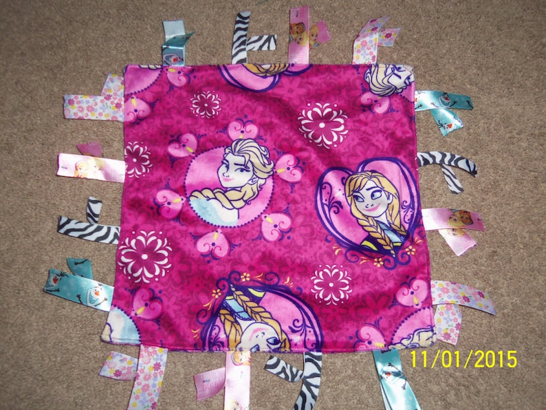 Peut inclure: Une couverture pour b&eacute;b&eacute; en tissu rose et bleu sur le th&egrave;me de La Reine des Neiges. La couverture a un fond rose avec des fleurs et des c&oelig;urs blancs. Il y a trois images d'Elsa et d'Anna du film La Reine des Neiges. La couverture a plusieurs rubans et bandes de tissu attach&eacute;s aux bords.