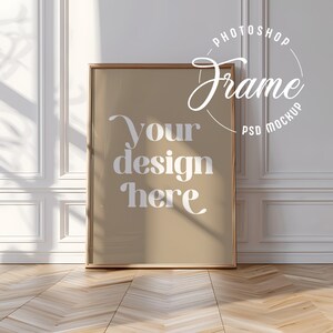 Könnte beinhalten: Ein rechteckiger gerahmter Poster-Mockup mit dem Text "your design here" in Weiß. Der Rahmen hat eine hellbraune Holzoberfläche. Das Poster lehnt an einer weißen Wand mit dekorativen Leisten, und der Boden ist aus hellem Holz.