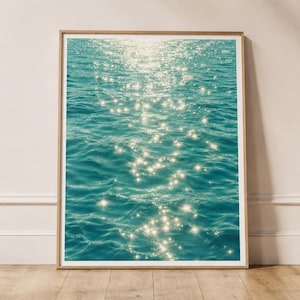 Puede incluir: Una impresión de arte mural rectangular que presenta una fotografía de agua de mar de color turquesa profundo con luz solar blanca brillante que resplandece sobre las ondas. La impresión está enmarcada en un marco de madera clara y sencilla.