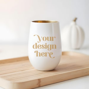 Puede incluir: Una copa de vino sin tallo blanca con un borde dorado y el texto "Your design here" en letras doradas. La copa está sobre una bandeja de madera marrón claro, con una calabaza blanca en el fondo.