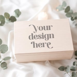 Könnte beinhalten: Eine hellfarbene Holzkiste mit dem Text "Your design here" auf dem Deckel. Die rechteckige Kiste liegt auf einer weißen, strukturierten Oberfläche, umgeben von Eukalyptuszweigen und brennenden Kerzen.