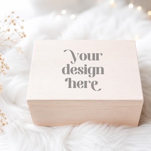 Könnte beinhalten: Eine hellfarbene Holzkiste mit dem Text "Your design here" auf der Oberseite. Die Kiste ist rechteckig und liegt auf einer weißen, flauschigen Oberfläche. Getrocknete Blumen befinden sich auf der linken Seite der Kiste.