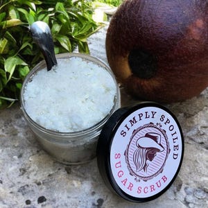 Peut inclure: Un pot en verre rempli de gommage au sucre blanc avec un couvercle noir sur lequel est inscrit "Simply Spoiled Sugar Scrub" en lettres blanches. Le pot est pos&eacute; sur une surface textur&eacute;e grise.