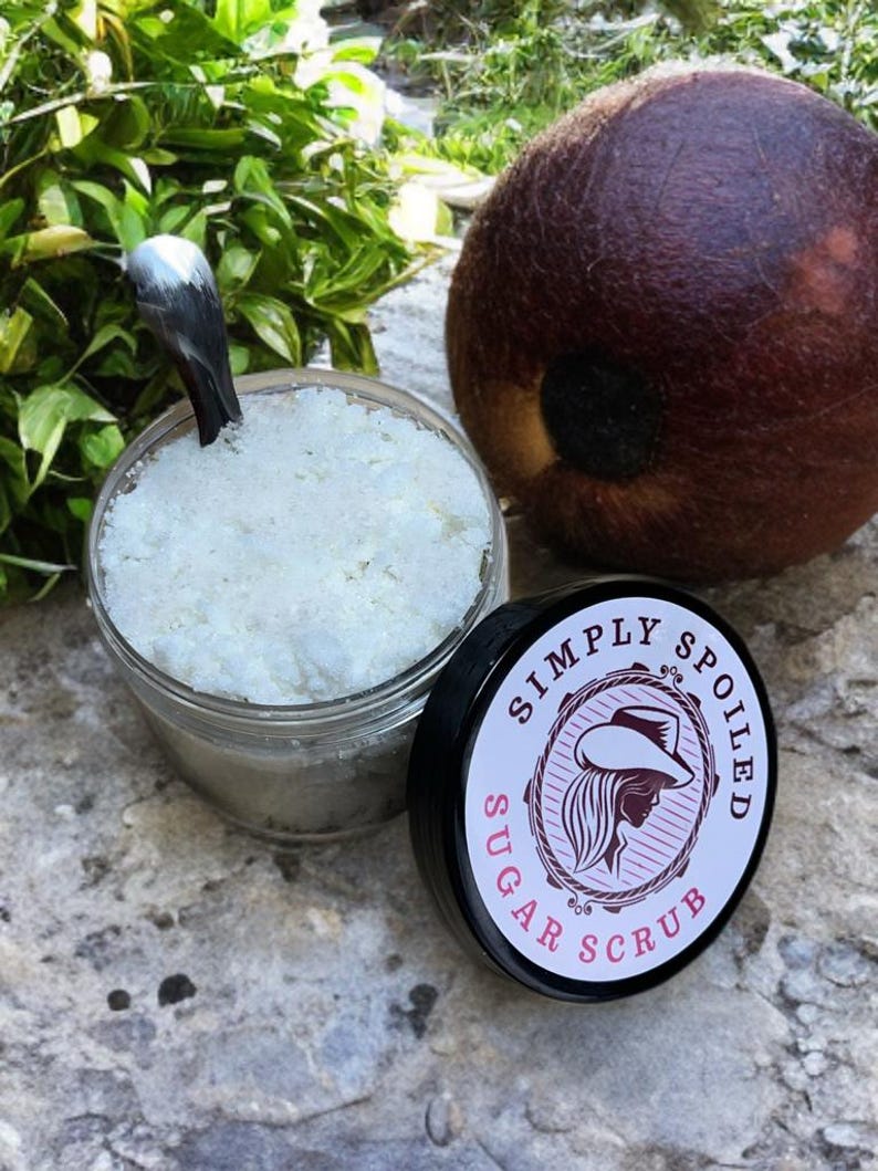 Peut inclure: Un pot en verre rempli de gommage au sucre blanc avec un couvercle noir sur lequel est inscrit "Simply Spoiled Sugar Scrub" en lettres blanches. Le pot est pos&eacute; sur une surface textur&eacute;e grise.