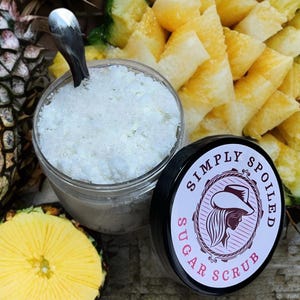 Peut inclure: Un pot en verre rempli de gommage au sucre blanc, avec un couvercle noir qui dit "Simply Spoiled Sugar Scrub". Le pot est entour&eacute; de tranches d'ananas.