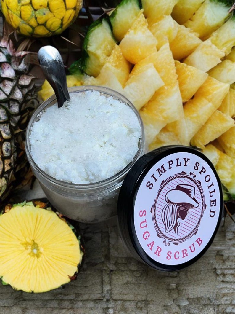 Peut inclure: Un pot en verre rempli de gommage au sucre blanc, avec un couvercle noir qui dit "Simply Spoiled Sugar Scrub". Le pot est entour&eacute; de tranches d'ananas.