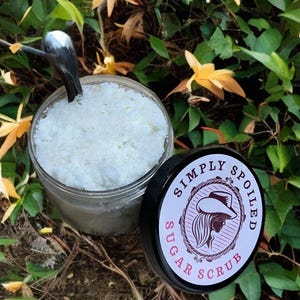 Peut inclure: Un pot en verre rempli de gommage au sucre blanc, avec un couvercle noir sur lequel est inscrit "Simply Spoiled Sugar Scrub" en lettres blanches avec une silhouette rose d'une femme portant un chapeau de cow-boy.