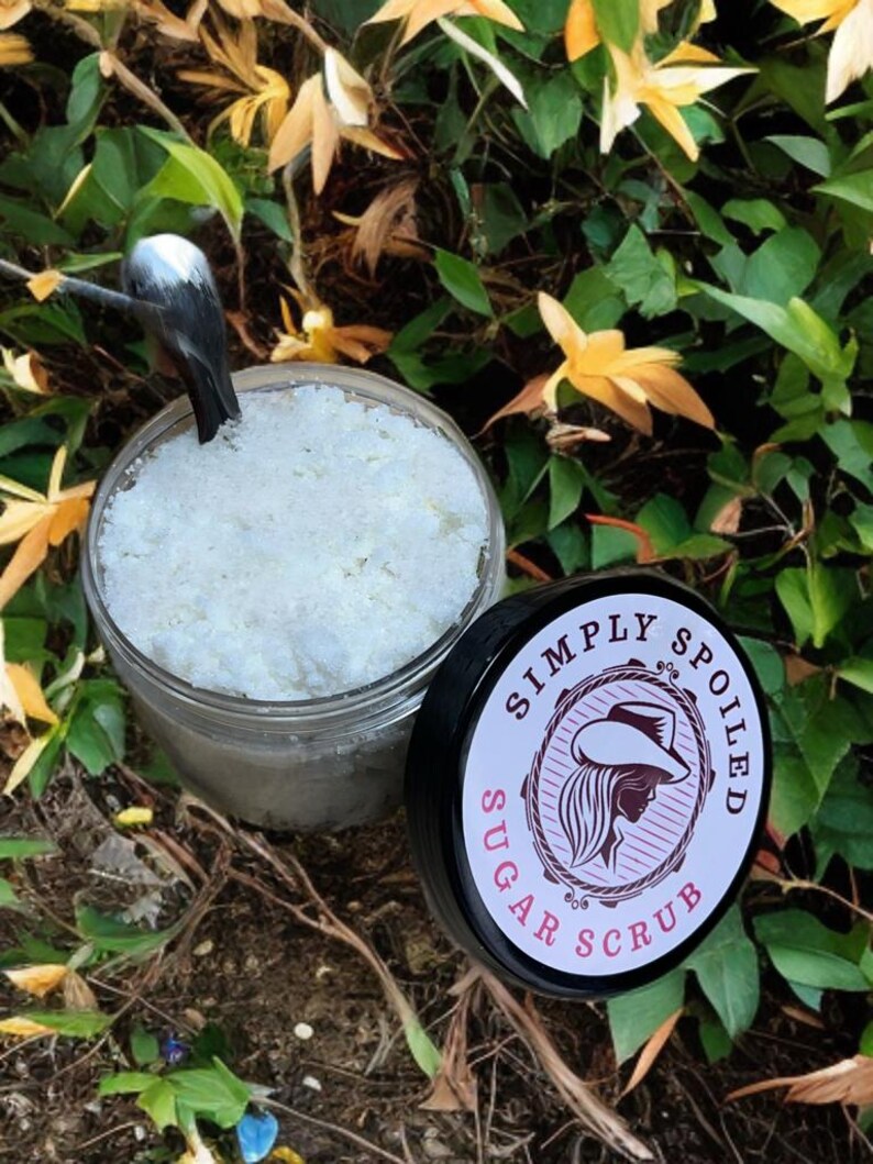 Peut inclure: Un pot en verre rempli de gommage au sucre blanc, avec un couvercle noir sur lequel est inscrit "Simply Spoiled Sugar Scrub" en lettres blanches avec une silhouette rose d'une femme portant un chapeau de cow-boy.