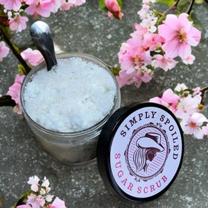 Peut inclure: Un pot en verre rempli de gommage au sucre blanc, avec un couvercle noir qui dit "Simply Spoiled Sugar Scrub". Le pot est pos&eacute; sur une surface grise avec des fleurs roses.