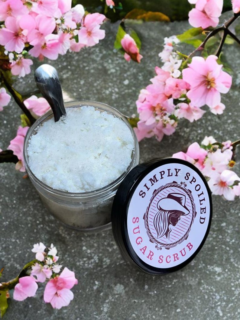 Peut inclure: Un pot en verre rempli de gommage au sucre blanc, avec un couvercle noir qui dit "Simply Spoiled Sugar Scrub". Le pot est pos&eacute; sur une surface grise avec des fleurs roses.