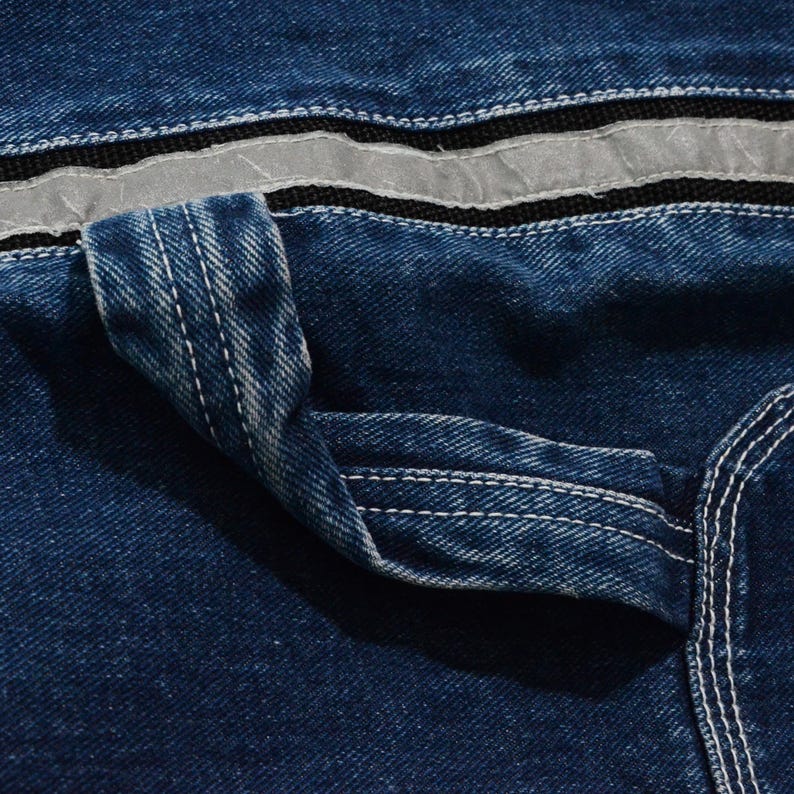 May include: Close-up of blue denim fabric with white stitching. Features a pocket detail and a reflective silver stripe. The denim has a classic, textured appearance, showcasing the sturdy construction of the garment.