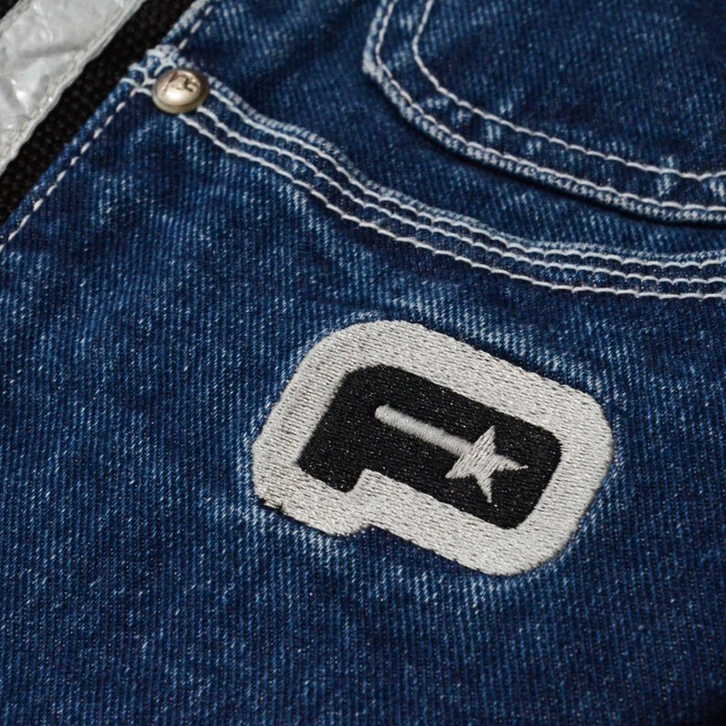 May include: Close-up of blue denim jeans with white stitching and a silver stripe. The jeans feature a black and gray embroidered patch with a stylized letter and star design. A silver button is visible.