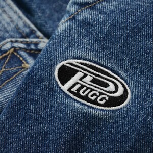 May include: Close-up of blue denim fabric with a white and black embroidered patch. The patch has the word "LUGG" in white lettering and a stylized "P" design.