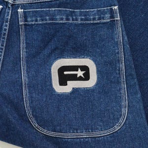May include: Close-up of a section of blue denim jeans. The back pocket features a gray patch with a black stylized letter "P" and a star. The stitching on the pocket and seams is visible.