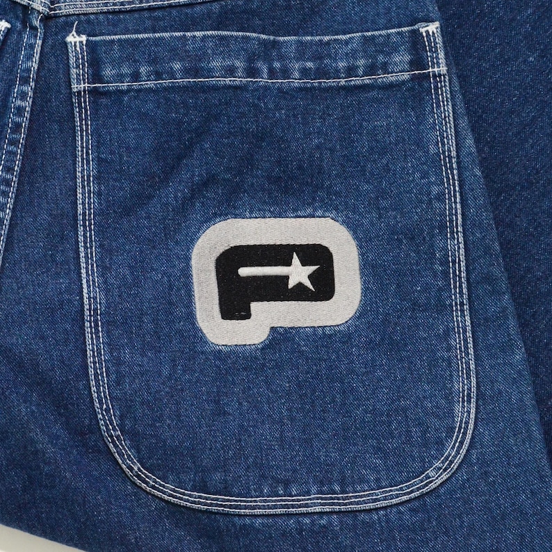 May include: Close-up of a section of blue denim jeans. The back pocket features a gray patch with a black stylized letter "P" and a star. The stitching on the pocket and seams is visible.