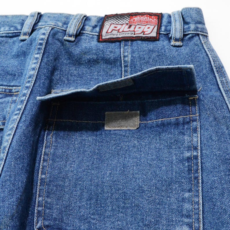 May include: Close-up of blue denim jeans with a back pocket and a rectangular patch. The jeans have a "PLUGG Clothing Company" label on the back. The denim is a medium wash with visible stitching.