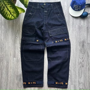 May include: Dark blue denim cargo trousers with multiple pockets and straps. The trousers feature yellow stitching and the text "M + FG" in yellow. A blue baseball cap with "POLO" text and headphones are in the background.